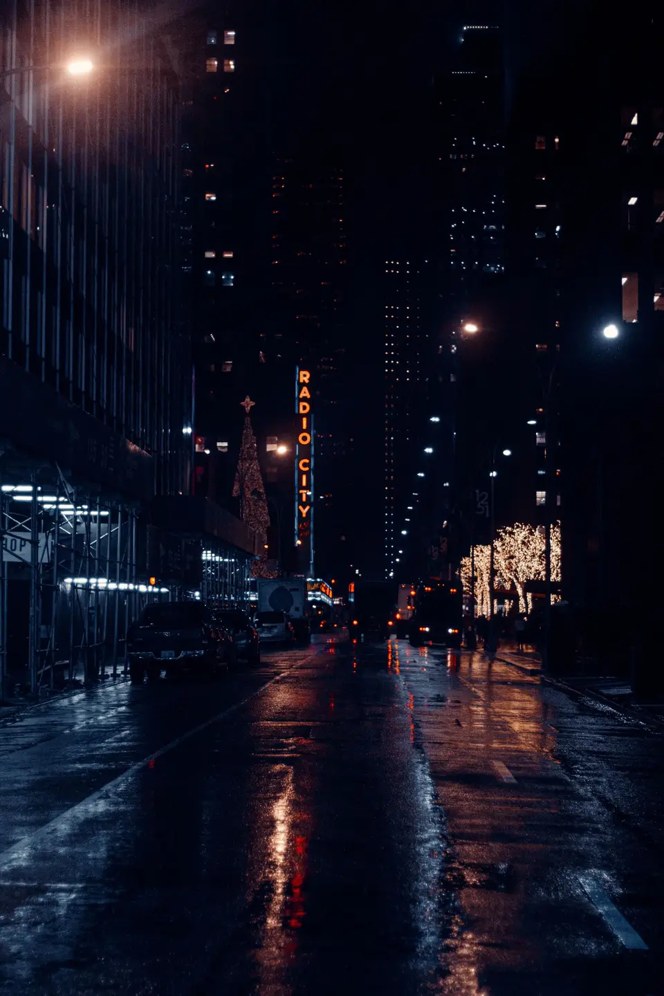 An empty, rain-wet Manhattan street at night, lit by street lamps, with the Radio City Music Hall sign small in the distance.